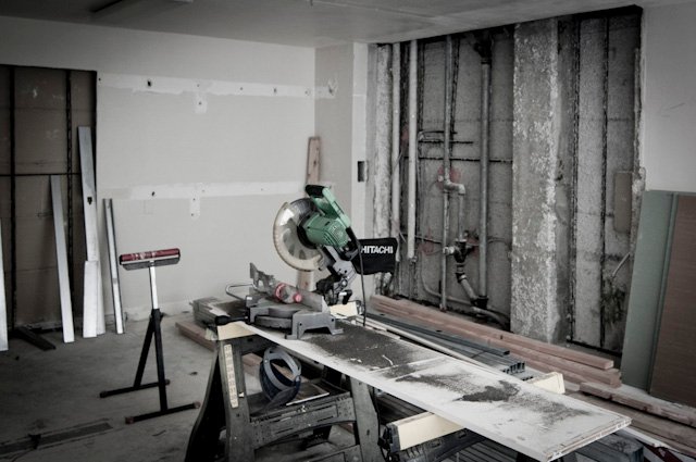 Room under renovation with exposed wall framing, pipes, lumber, and a miter saw