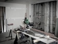 Room under renovation with exposed wall framing, pipes, lumber, and a miter saw