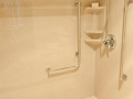 Beige walk-in shower with tiled floor, handheld showerhead, grab bars, and corner shelves