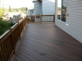 Spacious brown wood deck with railing extending along the side of a gray house