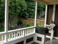 Covered porch with white railings, brown panels, and a view of a leafy backyard