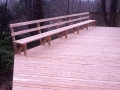Large unfinished wood deck with a long built-in bench and railing surrounded by trees