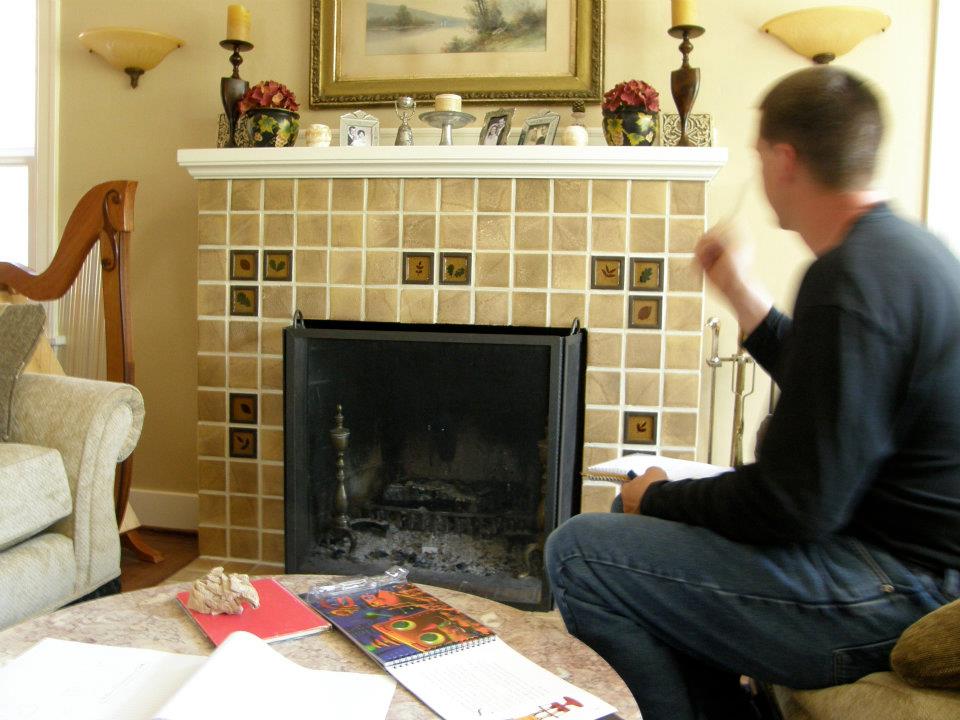 Man seated beside a beige tiled fireplace holding papers and gesturing