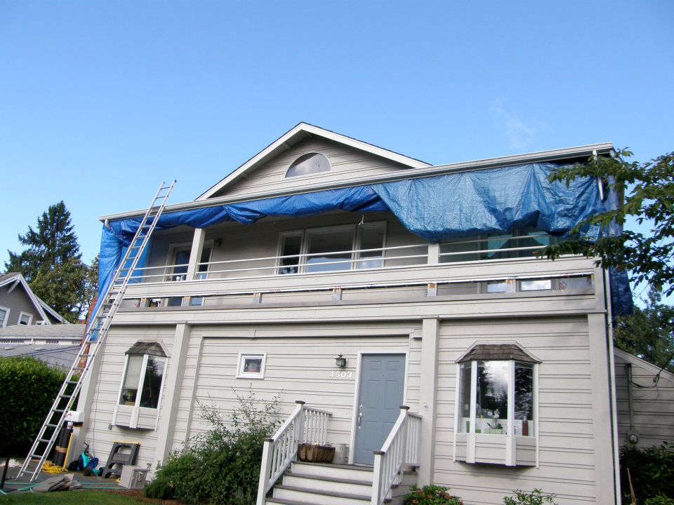 Two-story gray house with blue tarps and a ladder during exterior renovation
