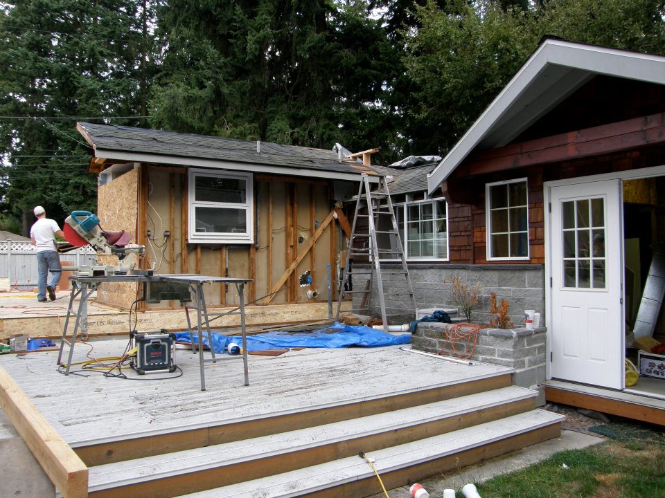 Home exterior under renovation with exposed framing, deck, ladders, tools, and worker