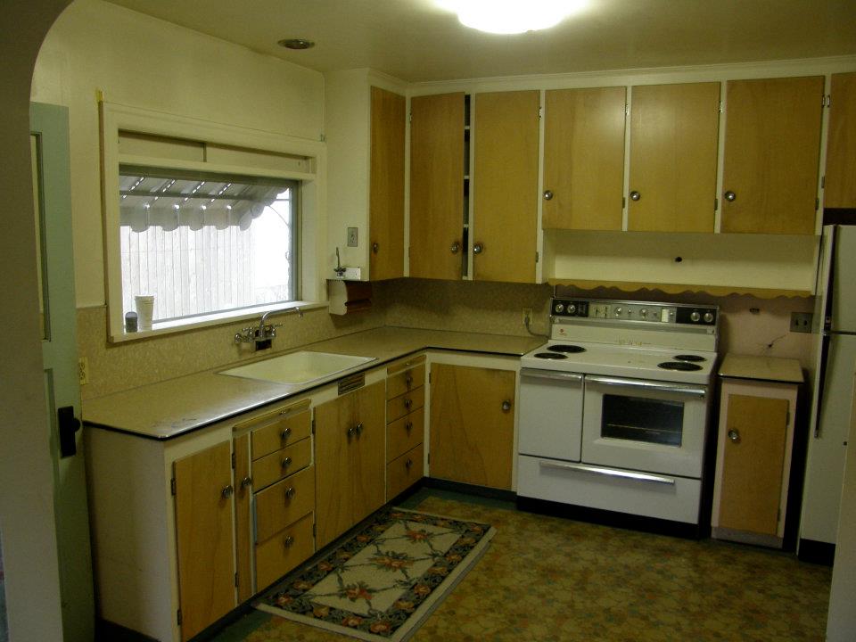 Older kitchen with wood cabinets, beige counters, white stove, and sink beneath a window