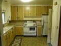 Older kitchen with wood cabinets, white stove and refrigerator, and beige countertops